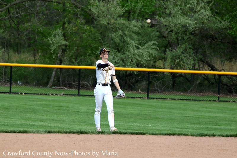Baseball player in a white uniform stands on the grass near the infield, glove up, as a ball flies overhead toward the outfield fence.