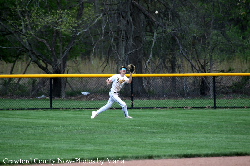 Baseball fielder in white uniform reaching up with glove to catch a fly ball along the outfield fence on a grassy field.