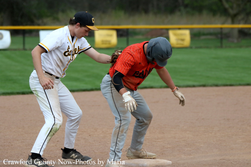 Two youth baseball players near a base: a fielder in white reaches toward the runner in orange on the dirt infield near the base.