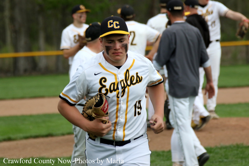 Baseball player in a white Eagles uniform with number 19 runs on the field, holding a glove, with black eye paint under his eyes; teammates are blurred in the background.