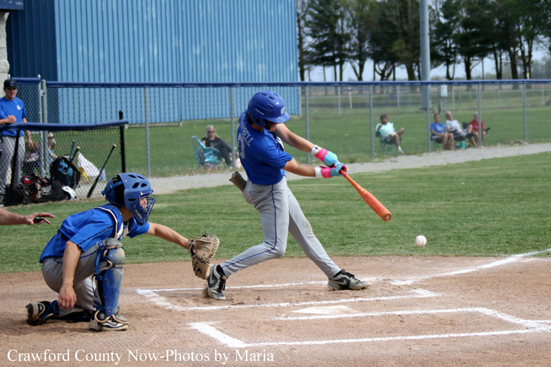Youth baseball action: a batter in a blue helmet and gray pants swings at a pitch while the catcher in blue crouches behind home plate on a dirt infield, ball in midair near the plate; spectators sit beyond the chain-link fence in the background.