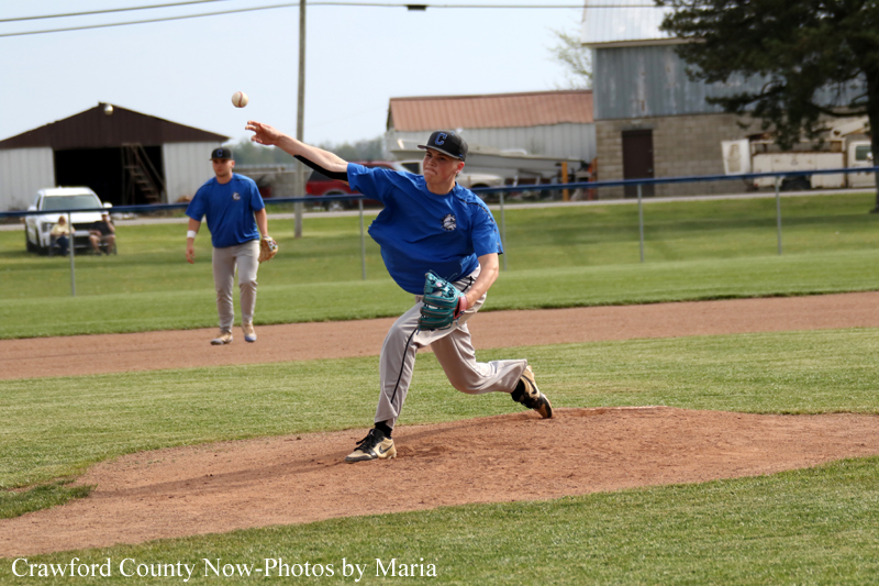 A baseball pitcher in a blue jersey winds up to throw from the mound as a fielder watches from behind on the field.