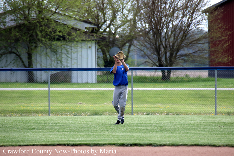 Young baseball player in a blue jersey and gray pants catches a fly ball with a mitt on a grassy field behind a chain-link fence.
