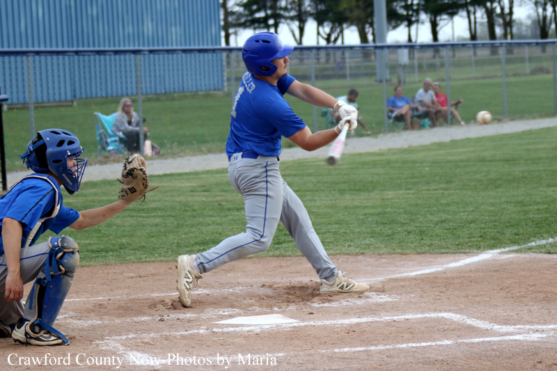 Baseball batter in a blue uniform swings at a pitch as the catcher in blue gear crouches behind him on the dirt at home plate, with spectators in the backgroundRe