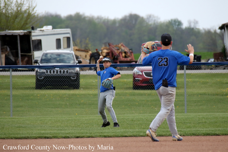 Young baseball pitcher in blue jersey throws a pitch on a grassy field as an older player wearing #23 watches from the right.