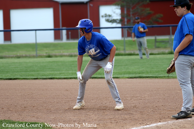 Youth baseball infield: a player in a blue helmet and jersey crouches, ready to field the ball with a glove.
