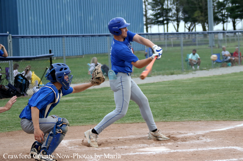 A baseball batter in a blue uniform swings at a pitch as the catcher in blue crouches behind home plate on a dusty infield with spectators in the background.
