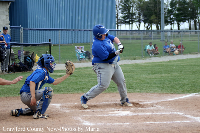 Youth baseball batter in blue swings at the ball, with a catcher crouched behind home plate and dirt infield nearby.