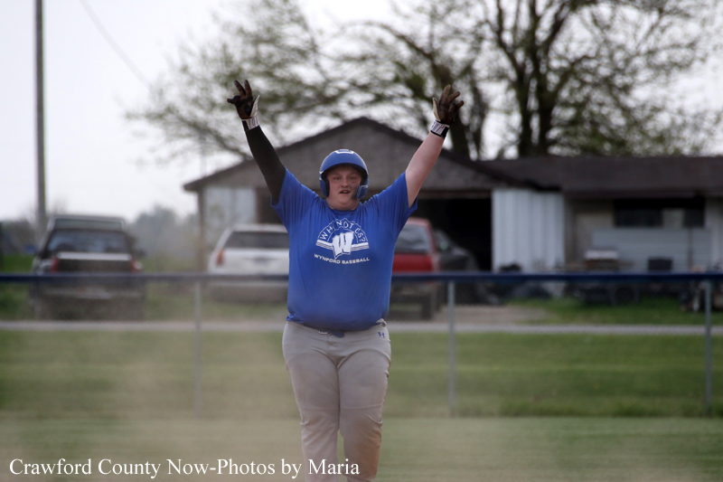 Softball player in a blue jersey and helmet raises arms in celebration on a field with a fence and buildings in the background.