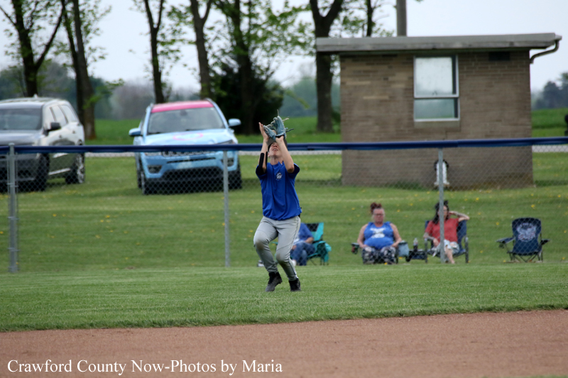 Baseball player in a blue shirt reaches up to catch a ball with a glove on a grassy field, spectators seated behind a chain-link fence in the background.