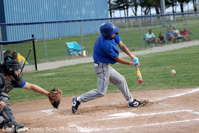 Boy in blue batting at a baseball game, swing about to hit the ball near home plate with catcher and spectators in the background.