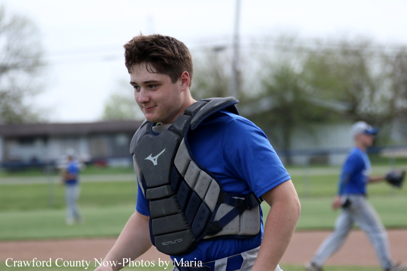 Baseball catcher in blue shirt wearing chest protector walks on the dirt infield with teammates in the background.