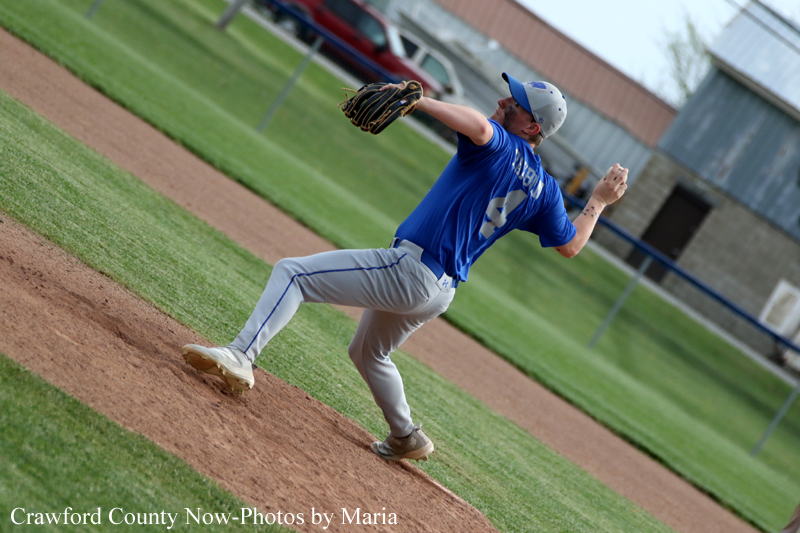 Baseball pitcher in a blue jersey throws from the mound during a game, left leg lifted in a pitching stance on a dirt mound.