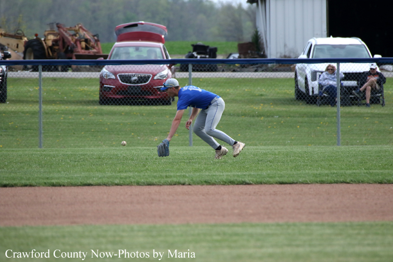 Baseball fielder in a blue shirt and gray pants lunges to field a ground ball on the grass, with a chain-link fence and parked cars in the background.