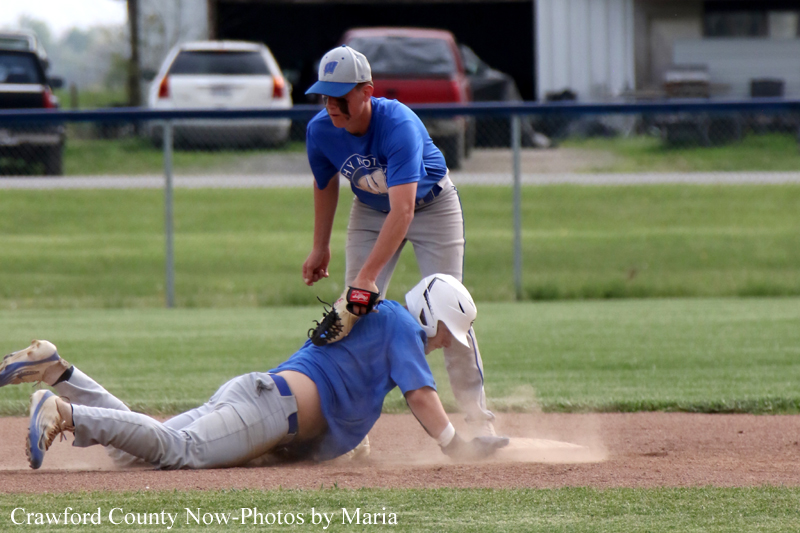Baseball player slides headfirst into a base as a teammate reaches with a glove to tag him. Dust rises from the infield dirt as the play unfolds.