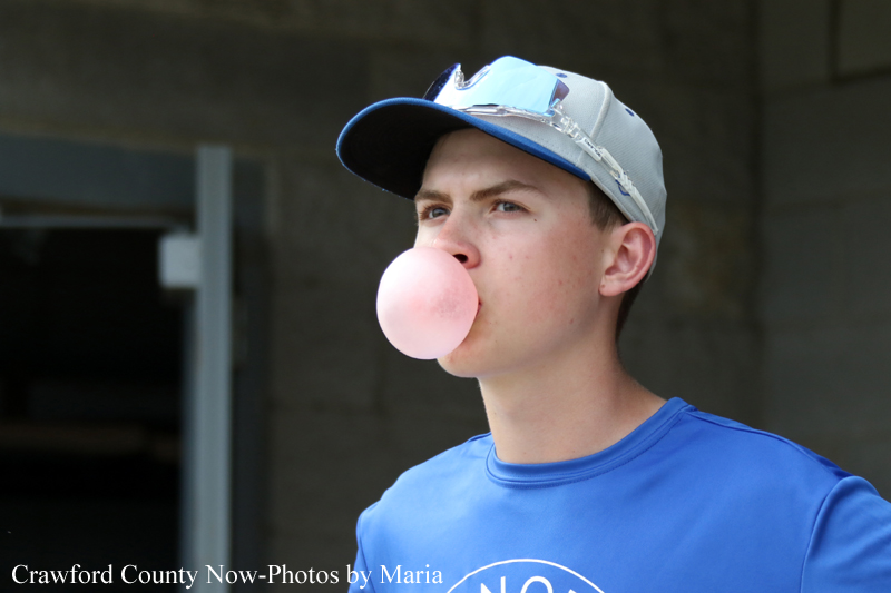 Teen wearing a blue shirt and cap blows a pink bubble gum outdoors, looking to the side.