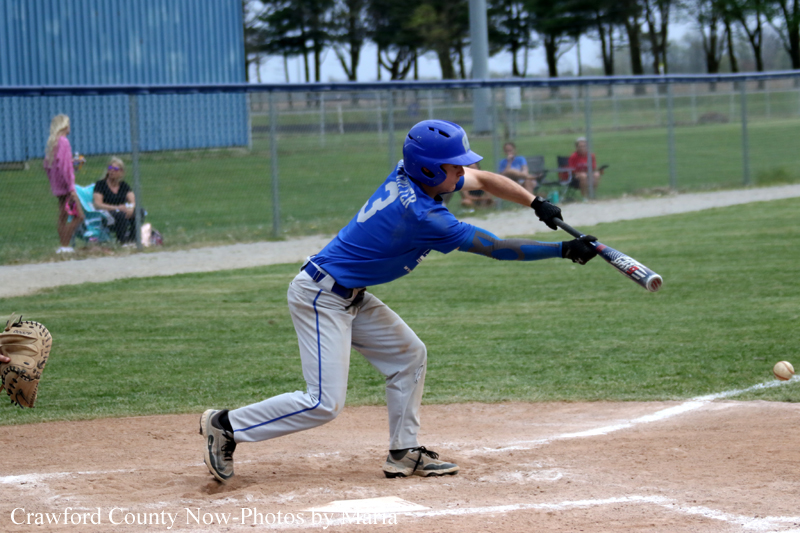 Baseball batter in a blue uniform swings at a ball near home plate as spectators watch in the background.