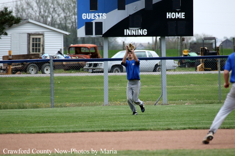 Baseball outfielder in a blue jersey reaches up to catch a fly ball in the outfield, with a chain-link fence, scoreboard, and parked vehicles in the background.