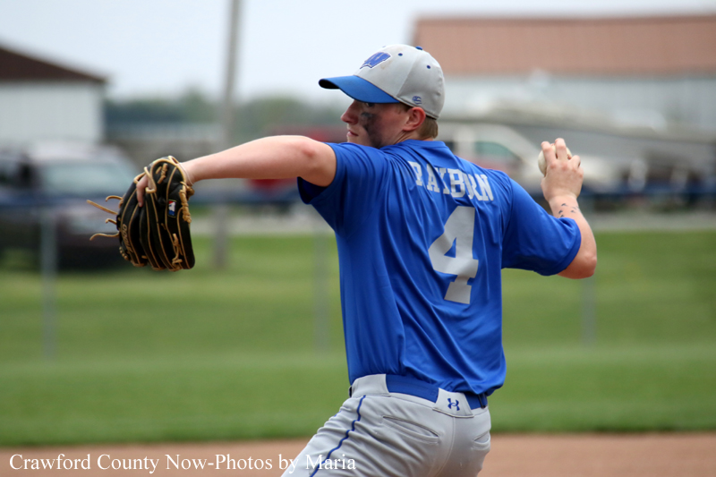 Pitcher in a blue jersey and gray cap throwing a pitch on a baseball field, glove outstretched to the left.