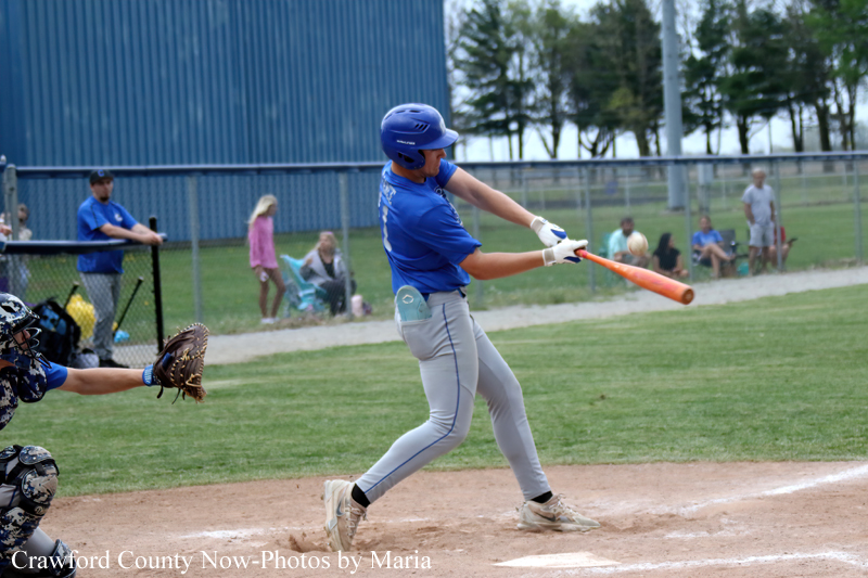 A baseball batter in blue swings at an incoming pitch as a catcher reaches behind him at home plate; spectators sit along the fence in the background. Washed-out field and blue building visible.