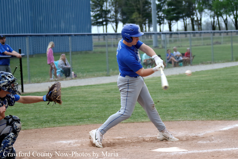 Baseball batter in a blue uniform swinging at a pitched ball at home plate, with a catcher in a glove behind him and a chain-link fence in the background.