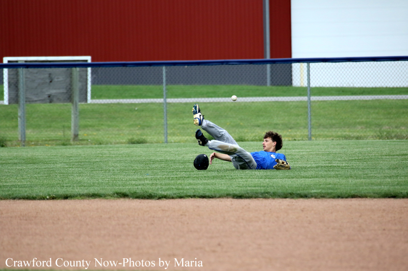 Baseball player slides on his back across the grass as a ball sails overhead, with a helmet and glove nearby.