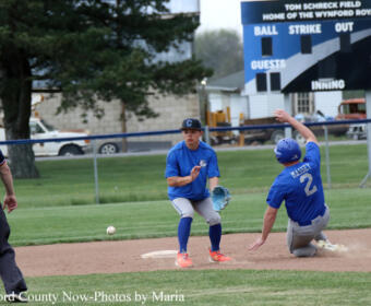 Baseball action: a fielder in blue prepares to tag a sliding runner near a base, with a scoreboard in the background.