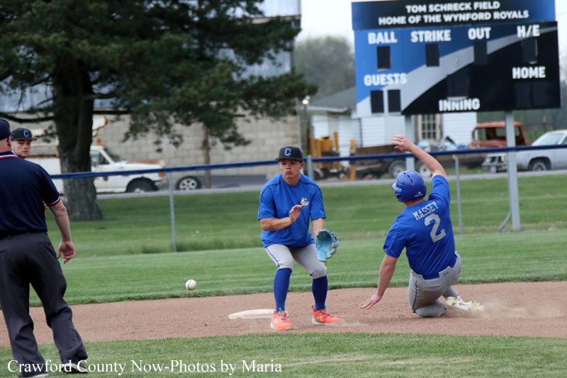 Baseball action: a fielder in blue prepares to tag a sliding runner near a base, with a scoreboard in the background.