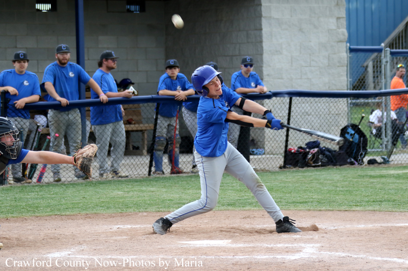 Young baseball batter in a blue jersey swings at a pitch as the ball sails toward him, with a catcher on the left and teammates watching from the dugout behind a chain-link fence.