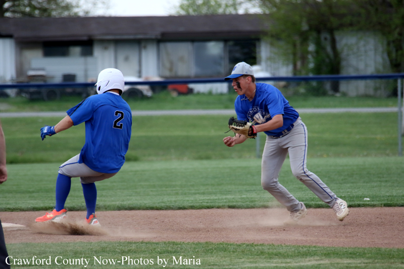 Two baseball players in blue uniforms on a dirt infield; a baserunner slides into a base while a fielder advances toward the play.