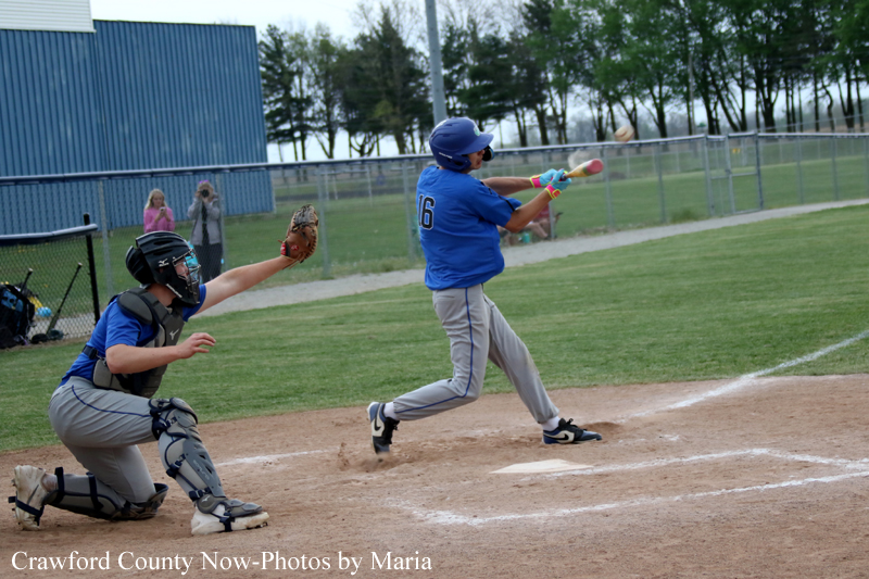Young baseball batter in a blue jersey swings at a pitch while the catcher in gray protective gear crouches behind him on the dirt infield.