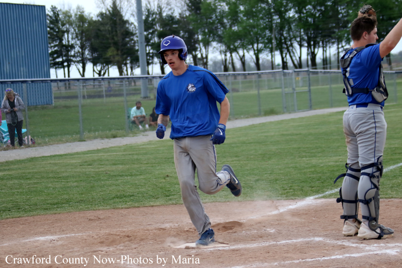 Baseball player in blue runs on the dirt infield during a game, with a catcher in blue and protective gear nearby on the right and spectators beyond the fence.