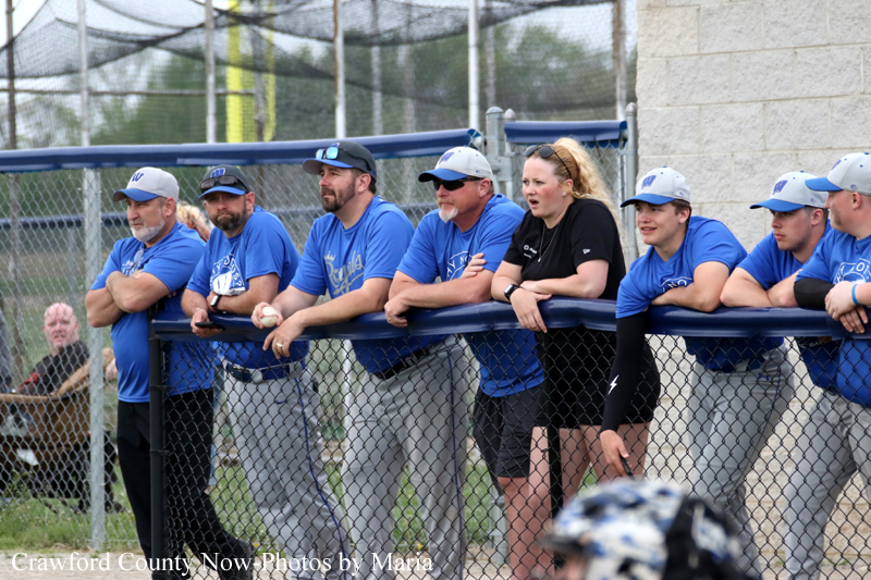 Line of baseball players in blue shirts and caps, plus a woman in black, leaning on the dugout railing and watching a game.