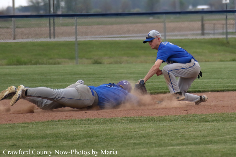Two baseball players in blue uniforms collide at a base: one sliding headfirst into the dirt as dust rises, the other crouched nearby ready to tag