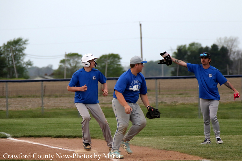 Three baseball players in blue shirts on a dirt infield during a game or drill; a runner wearing a white helmet moves toward a base as another teammate crouches, and a third player on the right raises his glove and hands toward the play.