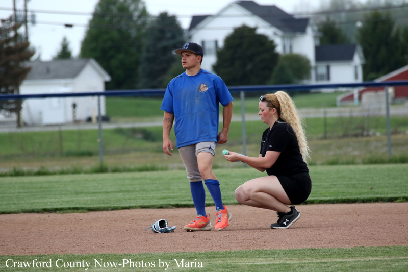 A baseball or softball player in a blue uniform standing on the infield while a coach kneels beside him, presenting or examining a ball on a dirt field with a helmet on the ground nearby.