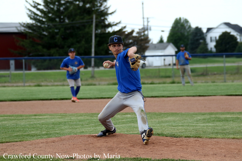 Right-handed pitcher in blue uniform winds up on the mound, preparing to throw, with teammates in the background.