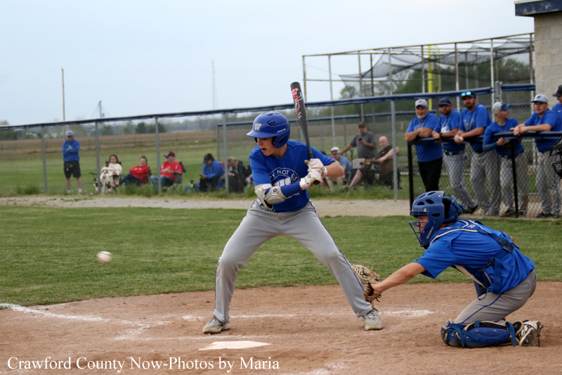 Baseball batter in a blue uniform swings at a pitch, with the catcher in blue gear ready behind him at home plate.