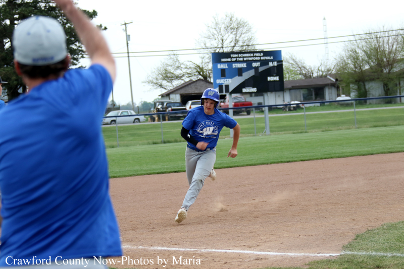 Baseball player in blue uniform sprinting on the dirt infield toward first base, with a teammate in blue in the foreground raising an arm.