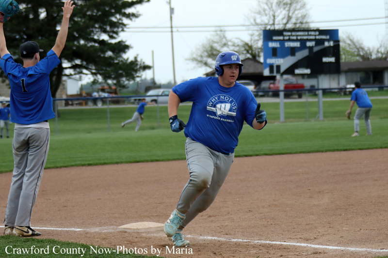 Baseball player in blue with helmet running toward first base as another fielder raises his arms; scoreboard and outfield in background.