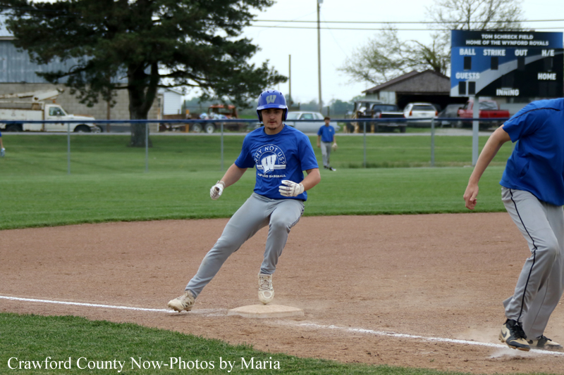 Baseball player in a blue uniform slides into second base during a game, with teammates and a scoreboard in the background.