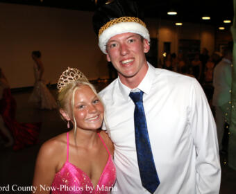 Two teens posing at a formal dance: girl in a pink sequined dress with a tiara, boy in a white shirt, blue tie, and a crown-like hat, smiling.