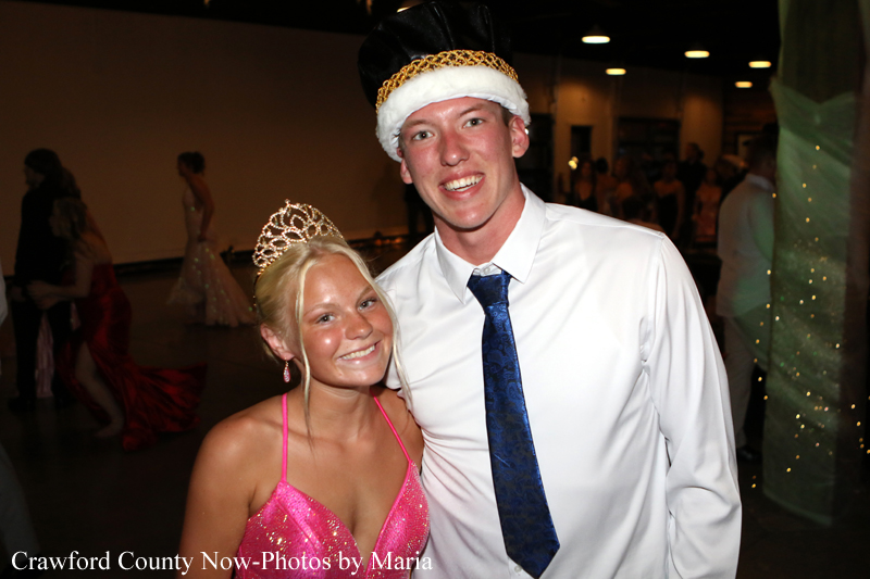 Two teens posing at a formal dance: girl in a pink sequined dress with a tiara, boy in a white shirt, blue tie, and a crown-like hat, smiling.