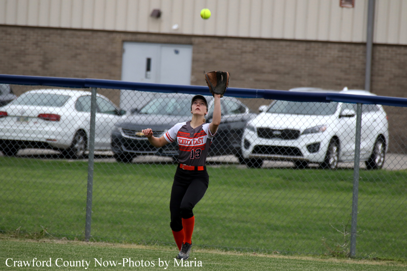 Female softball fielder in black and red uniform reaching up with her glove to catch a ball near the outfield fence.
