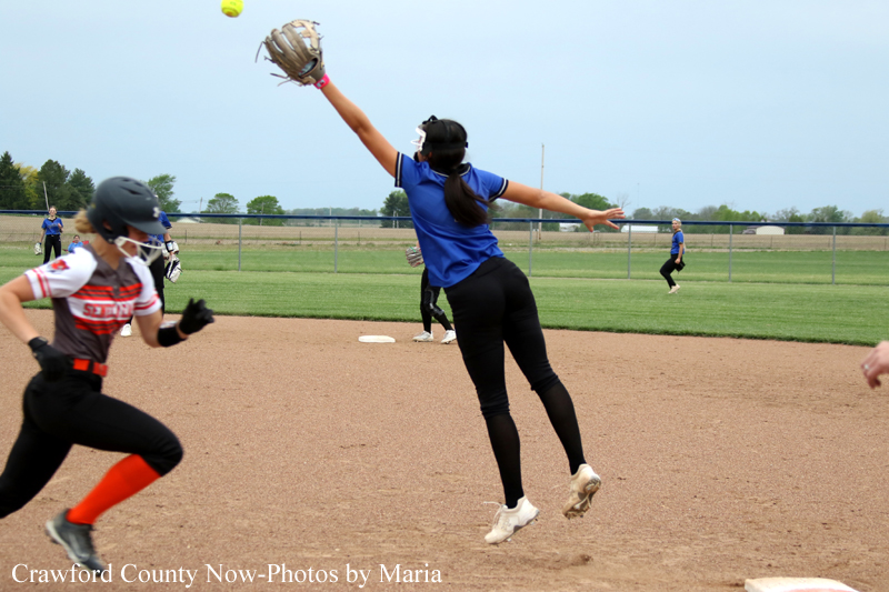 Female softball player in blue jumps to catch ball with glove raised on the infield, other players in the background.