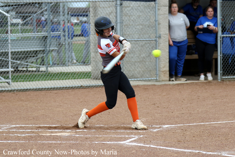 Young softball batter in a helmet swings at a pitch on the dirt field, wearing orange socks and a white-red uniform.