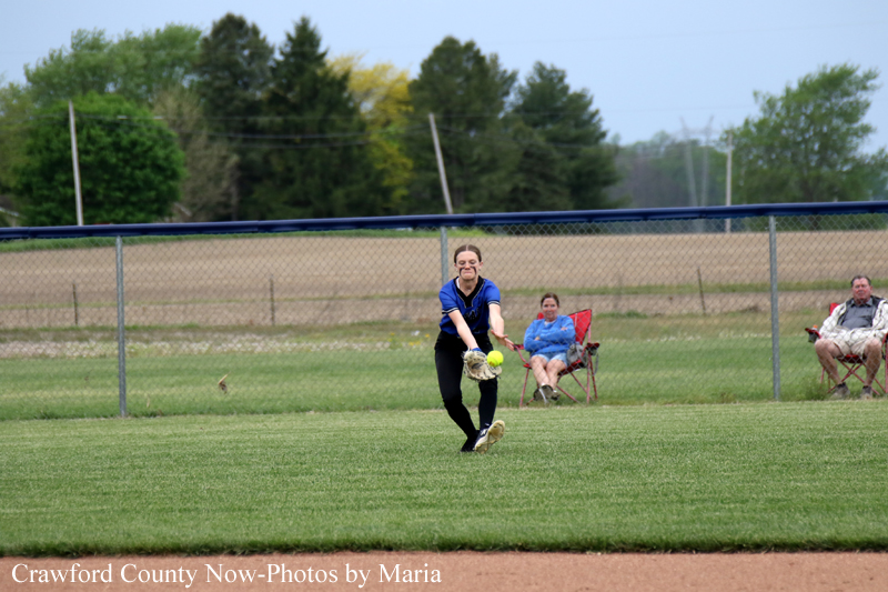 Young woman in a blue jersey pitches a softball on a grassy field, with two spectators seated behind a chain-link fence.
