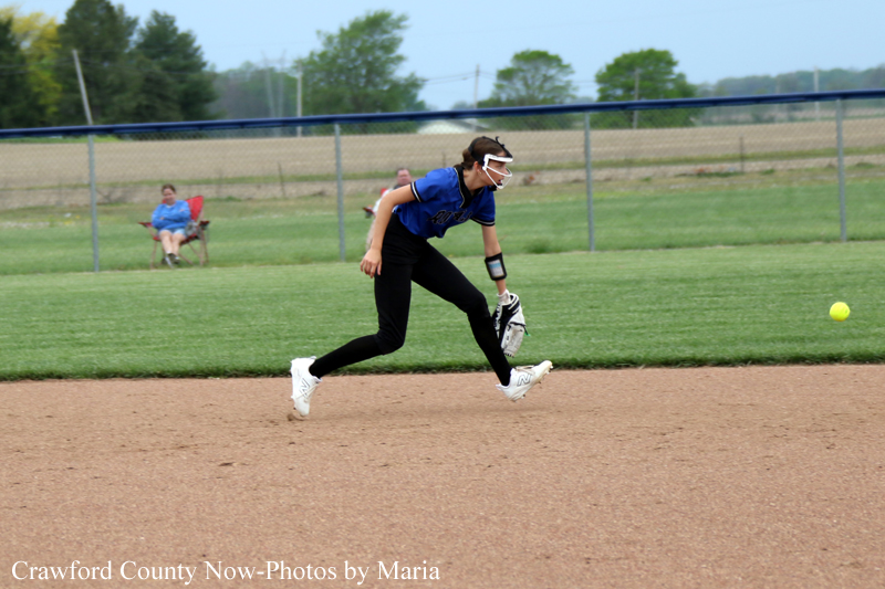 Female softball player in a blue jersey runs on the infield, glove ready, white visor on, as a yellow ball rolls to the right; spectator in the background.