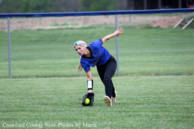 Female softball player in a blue Royals jersey dives to field a ground ball, glove near the grass on a sunny field.