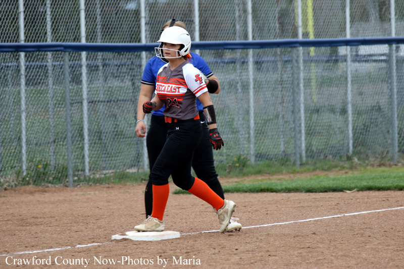 Softball player in a white helmet and orange socks runs toward first base on a dirt field, with a teammate behind and a chain-link fence in the background.